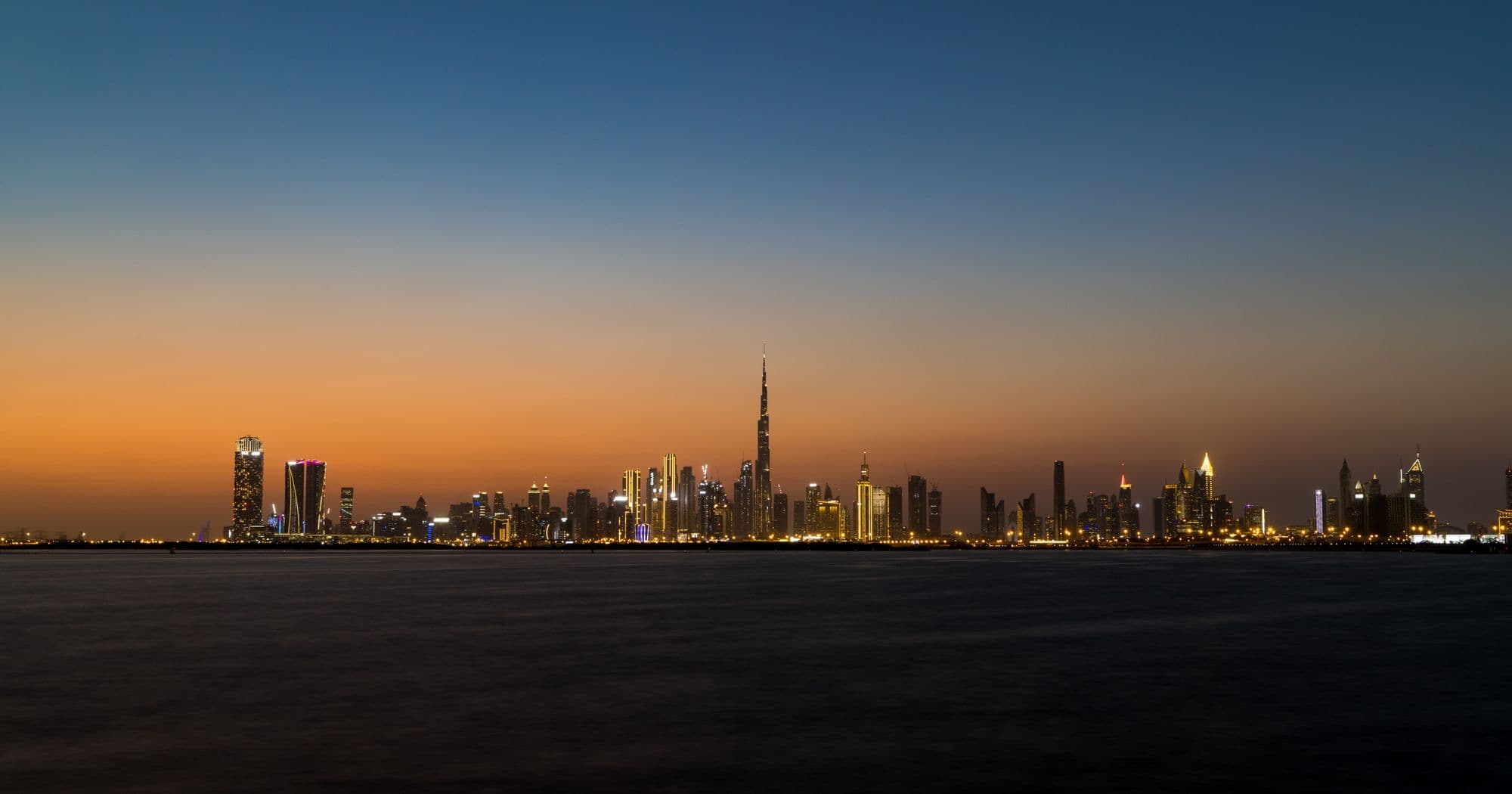 The Dubai skyline at dusk seen across the water, with the Burj Khalifa on the horizon